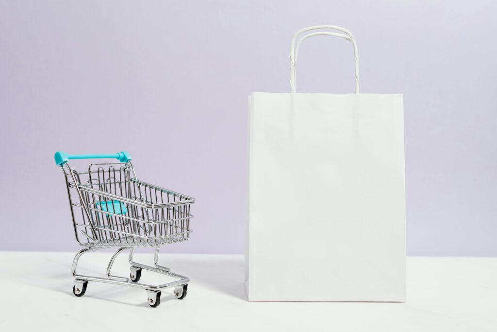 Minimalist image of a white shopping bag and mini cart on a soft pastel background.